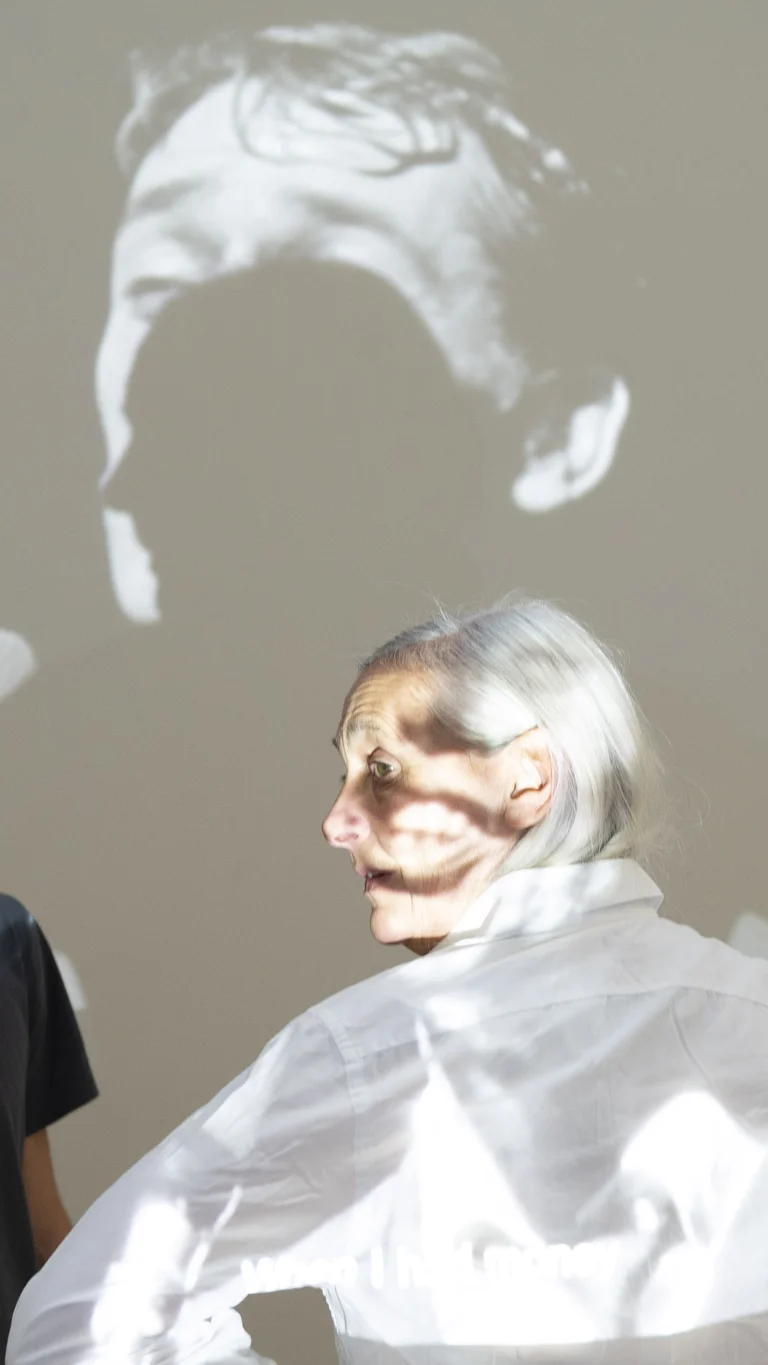 Solal Mariotte und Anne Teresa De Keersmaeker stehen vor einer hellen Wand, auf die ein schwarz-weißes Porträt projiziert wird. Beide blicken zur Seite. Anne Teresa De Keersmaeker trägt eine weiße Bluse und Solal Mariotte ein dunkles T-Shirt.