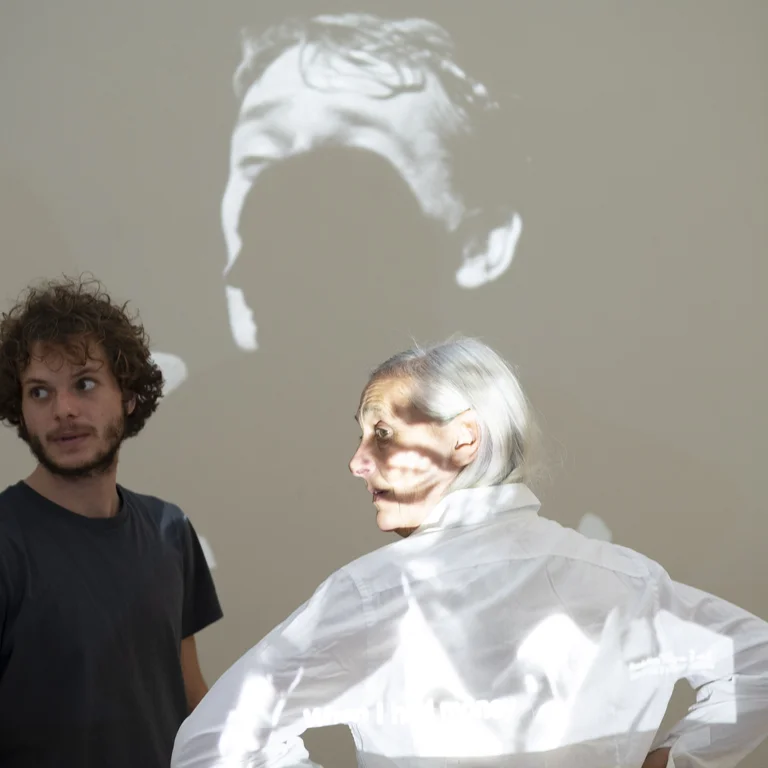 Solal Mariotte und Anne Teresa De Keersmaeker stehen vor einer hellen Wand, auf die ein schwarz-weißes Porträt projiziert wird. Beide blicken zur Seite. Anne Teresa De Keersmaeker trägt eine weiße Bluse und Solal Mariotte ein dunkles T-Shirt.