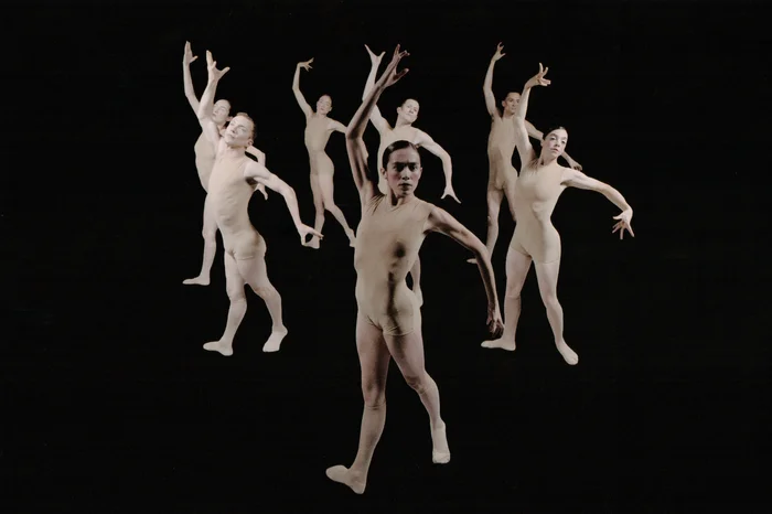 A group of dancers in skin-toned costumes move synchronously in expressive poses against a black background. The scene conveys harmony, energy, and collective presence.