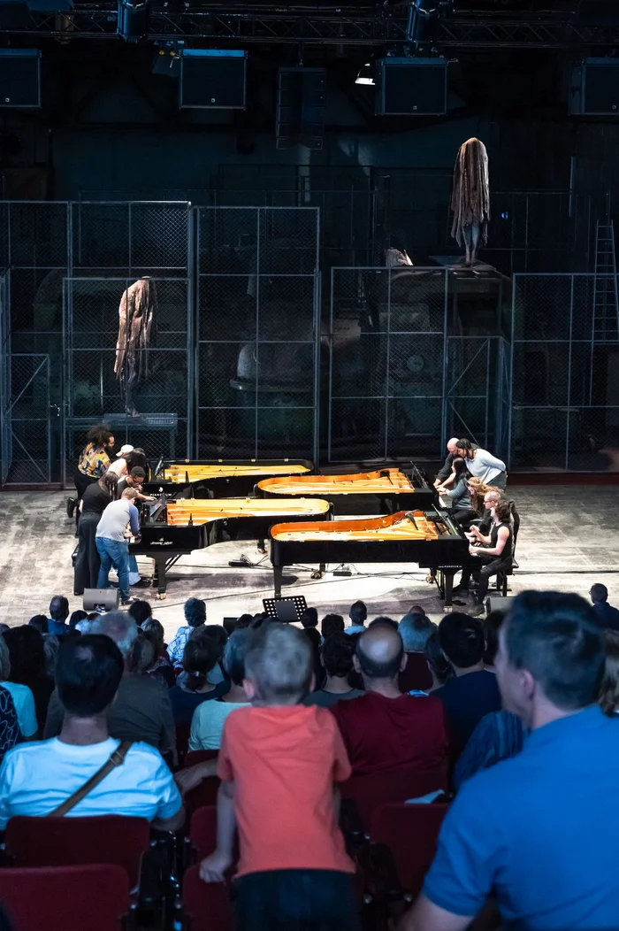 Das Bild zeigt das Appetizer-Konzert des Wild up Ensembles in der Turbinenhalle Bochum im Rahmen der Ruhrtriennale 2024. Mehrere Flügel sind auf der Bühne arrangiert, an denen die Musiker
intensiv arbeiten, um sie für das Konzert vorzubereiten. Im Hintergrund ist ein industriell anmutendes Bühnenbild zu sehen, das durch große Metallgitter und skulpturale Elemente dominiert wird. Das Publikum beobachtet gespannt die Vorbereitungen. Die Szene vermittelt eine besondere Verbindung zwischen dem rauen Industriecharakter der Halle und der klassischen Musikdarbietung.