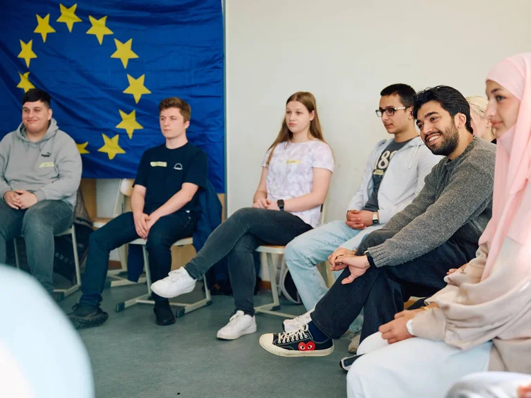 A group of young people sits in a circle of chairs in front of an EU flag, participating in a discussion or workshop.