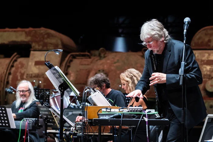 A musician from the Will Gregory Moog Ensemble stands in the foreground, focused on playing an analog synthesizer. In the background, other ensemble members are seen at their instruments, while the rusty turbines of the industrial hall provide a striking contrast to the modern electronic equipment.