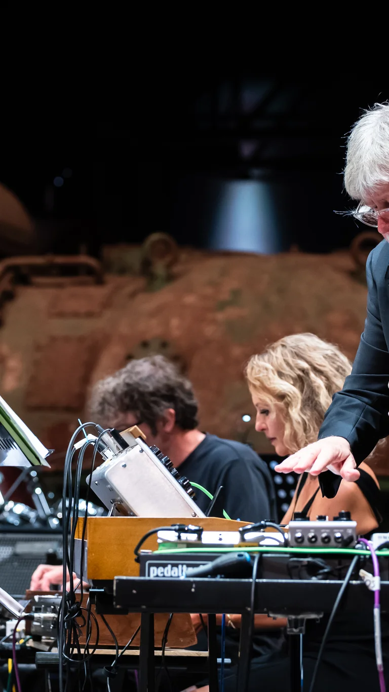 A musician from the Will Gregory Moog Ensemble stands in the foreground, focused on playing an analog synthesizer. In the background, other ensemble members are seen at their instruments, while the rusty turbines of the industrial hall provide a striking contrast to the modern electronic equipment.