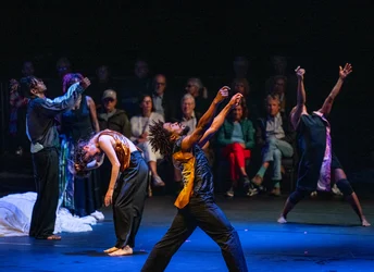 Several barefoot performers move across the stage with expressive, sweeping gestures. The audience sits directly behind the performance area, watching the dance performance.