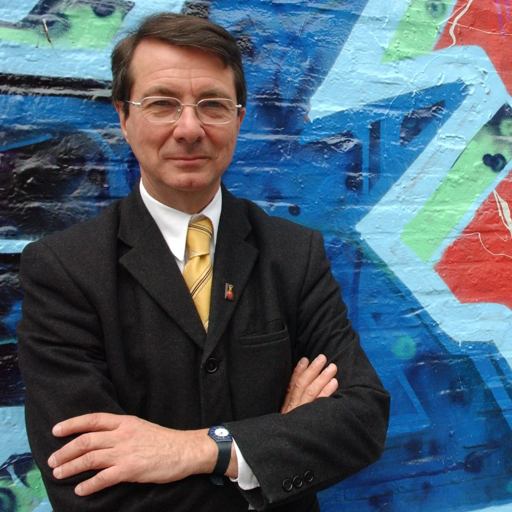 Gerard Mortier steht mit verschränkten Armen vor einer bunten Graffiti-Wand.