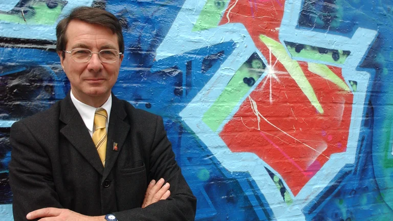 Gerard Mortier steht mit verschränkten Armen vor einer bunten Graffiti-Wand.