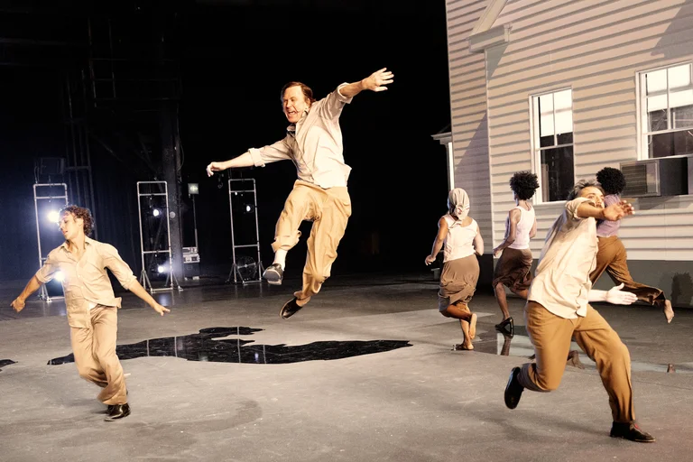 Several performers move dynamically across a theater stage. At the center, Lars Eidinger leaps into the air with outstretched arms and a joyful expression. Around him, other performers run and dance, dressed in beige and lilac-colored clothing. In the background, a white house facade with windows and a staircase is visible.