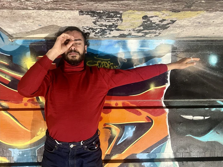 Mohamed Toukabri lies with his eyes closed and his right arm extended in front of a colorful graffiti wall. He wears a red turtleneck sweater and makes a circle gesture in front of one eye with his left hand.