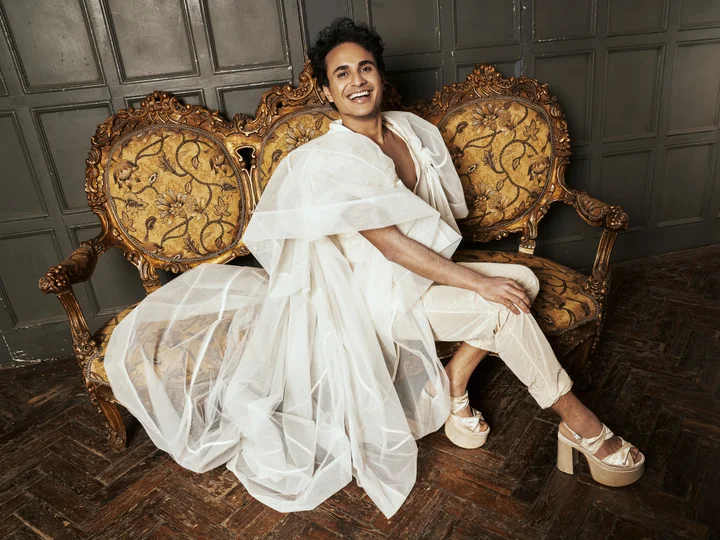 Samuel Mariño sits in elegant white flowing fabric on an ornate golden baroque sofa. He poses in a mix of classic and modern aesthetics against a dark-paneled wall.