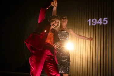Zwei Performerinnen in festlicher Kleidung tanzen in einer Szene aus "We Are the Lucky Ones", während eine Lichtquelle die Bühne erhellt.