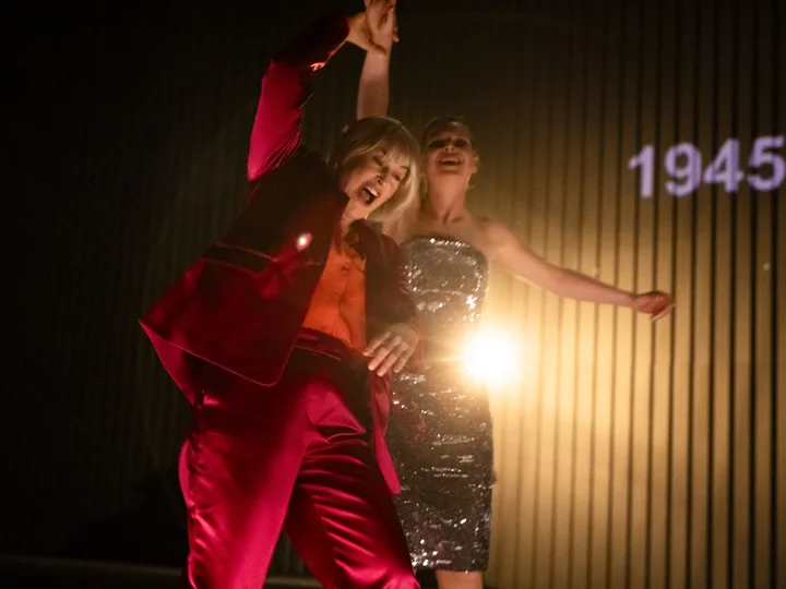 Zwei Performerinnen in festlicher Kleidung tanzen in einer Szene aus "We Are the Lucky Ones", während eine Lichtquelle die Bühne erhellt.