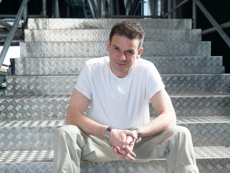 Łukasz Twarkowski is sitting on a staircase made of ribbed metal. He is wearing a white T-shirt, light brown trousers, and a wristwatch. His hands are folded in front of him.
