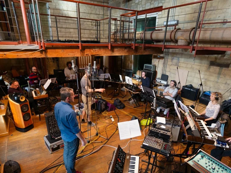Probenaufnahme des Will Gregory Moog Ensemble in einem Studio mit Steinwänden und Galerie. Die Musiker:innen sitzen in einem Kreis, umgeben von Synthesizern, Notenständern und Kabeln.