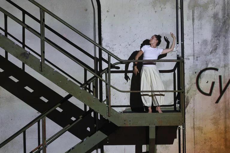Sarah Silverblatt-Buser stands on a metal staircase in an industrial building. She is wearing a white T-shirt and light-coloured trousers and raises one arm upwards. Light casts her shadow and the geometric shapes of the staircase onto the unfinished wall.