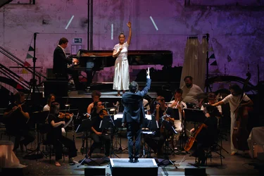 The ensemble Les Apaches ! performs on stage in an industrial hall, conducted by Julien Masmondet. In the background, Sarah Silverblatt-Buser stands with her arms raised in front of a grand piano, where a pianist is playing. The stage is bathed in purple light, with white lines of light projected onto the wall.