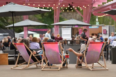 Das Bild zeigt das Festivalzentrum Wunderland bei der Ruhrtriennale 2024. Im Vordergrund entspannen sich drei Personen auf Liegestühlen, die mit dem Logo der Ruhrtriennale bedruckt sind. Im Hintergrund sind mehrere Besucher
zu sehen, die das gesellige Ambiente des Festivalzentrums genießen. Über ihnen hängen Lichterketten, die für eine stimmungsvolle Atmosphäre sorgen, während sich die Menschen um die verschiedenen Stände und Sitzmöglichkeiten verteilen.