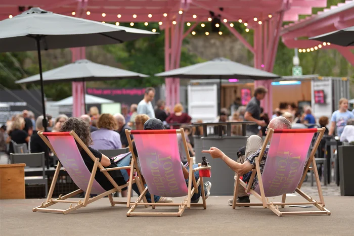 Das Bild zeigt das Festivalzentrum Wunderland bei der Ruhrtriennale 2024. Im Vordergrund entspannen sich drei Personen auf Liegestühlen, die mit dem Logo der Ruhrtriennale bedruckt sind. Im Hintergrund sind mehrere Besucher
zu sehen, die das gesellige Ambiente des Festivalzentrums genießen. Über ihnen hängen Lichterketten, die für eine stimmungsvolle Atmosphäre sorgen, während sich die Menschen um die verschiedenen Stände und Sitzmöglichkeiten verteilen.