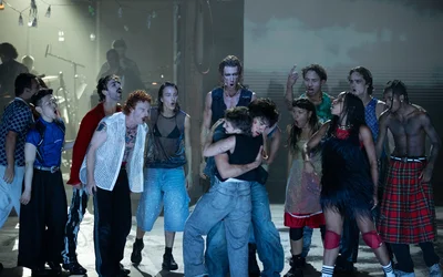 Performers stand close together on stage. The people in the center are embracing tightly. Those standing around them are glaring at them menacingly. Smoke is visible in the background.