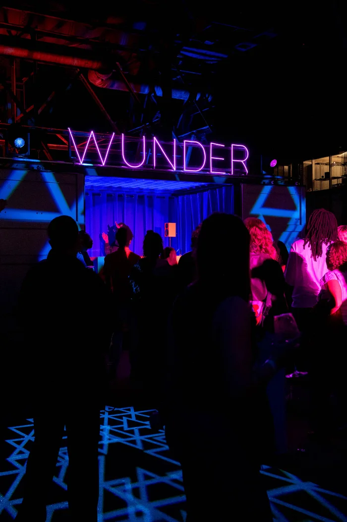 A crowd celebrates in front of the blue and pink-lit DJ area with an illuminated WUNDER sign.