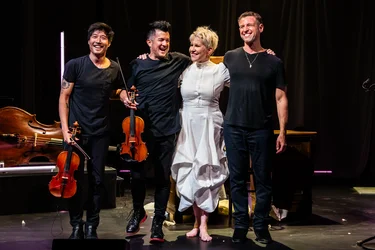 Charles Yang, Nick Kendall, Joyce DiDonato, and Ranaan Meyer stand smiling together on stage with their arms around one another. Joyce DiDonato wears a white dress, while the three musicians of Time for Three are dressed in black. Two violins and a double bass are visible beside them.