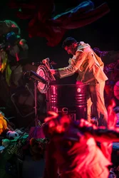 Performer schlägt mit Schlägeln auf ein Metallfass, das als Schlaginstrument aufgebaut ist; um ihn herum liegen Textilien und Alltagsobjekte auf der Bühne.