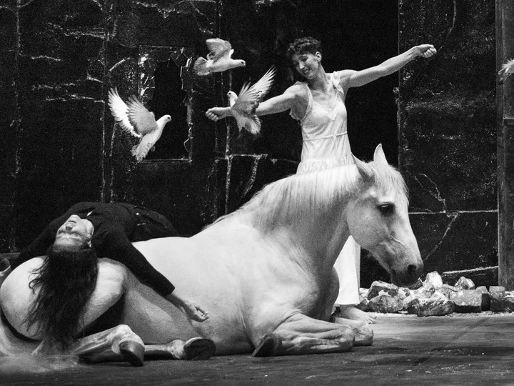 A performer in a light-coloured dress interacts with a white horse and flying doves on stage. Another person in dark clothing is lying on the horse. The photo is in black and white.