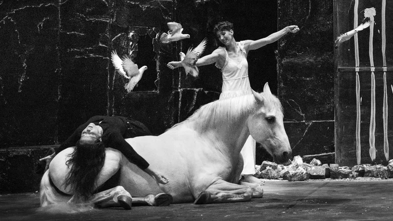 A performer in a light-coloured dress interacts with a white horse and flying doves on stage. Another person in dark clothing is lying on the horse. The photo is in black and white.