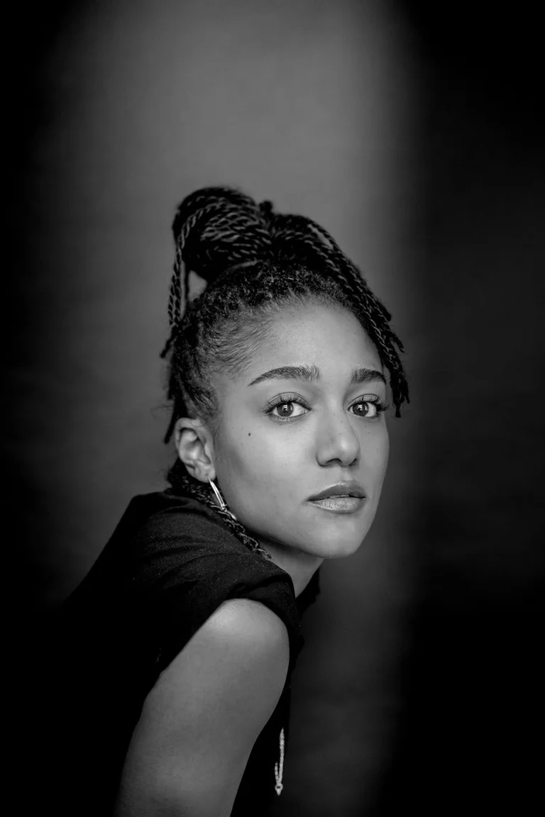 Ein Schwarz-Weiß-Porträt der Schauspielerin Larissa Sirah Herden. Sie blickt direkt in die Kamera. Ihr Haar ist nach oben gebunden, und sie trägt ein dunkles Oberteil und dezente Ohrringe. Sie sitzt vor einem dunklen, schlichten Hintergrund.