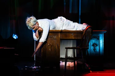 Joyce DiDonato lies on her stomach on a large wooden table, wearing a white dress. Her head rests on one arm while the other hangs down from the side of the table. A wooden chair stands beside the table on the dimly lit stage.