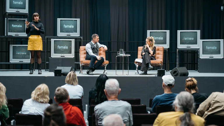 Auf einer Bühne sitzen Dr. Janosch Schobin und Sivan Ben Yishai in Sesseln im Gespräch, neben ihnen steht eine Frau und übersetzt in Gebärdensprache. Mehrere alte Fernseher im Hintergrund zeigen das Wort „female“. Im Vordergrund ist das Publikum zu sehen.