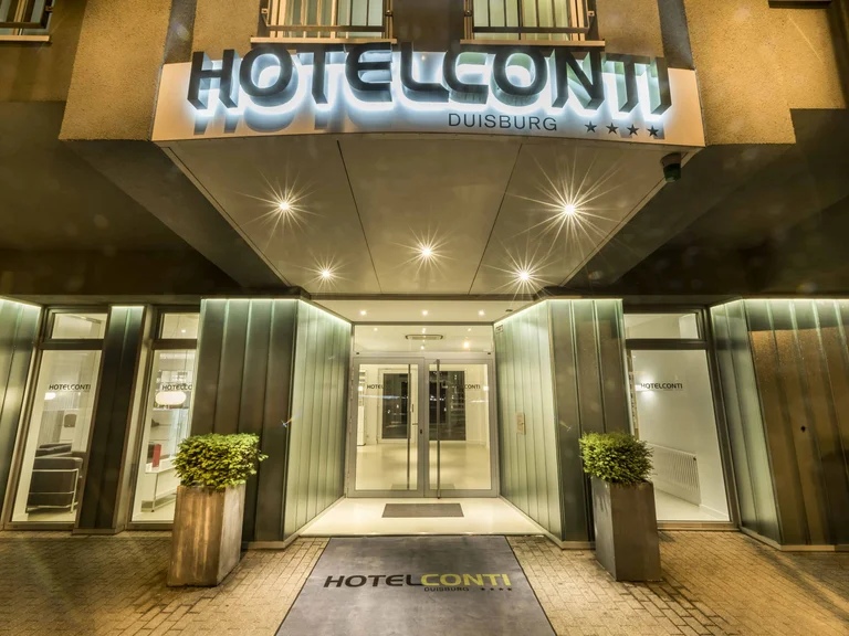Illuminated hotel entrance of Hotel Conti in Duisburg at night, with glass façade and planters.