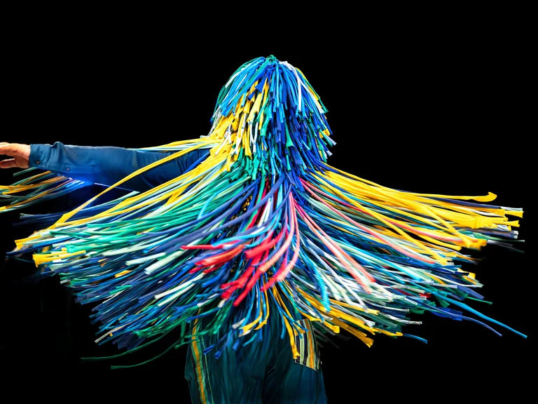 Eine Person in einem dunkelblauen Outfit bewegt sich mit ausgestrecktem Arm vor einem schwarzen Hintergrund. Ihr gesamter Kopf und Oberkörper sind mit langen, bunten Plastikstreifen bedeckt. Die Bewegung lässt die Streifen dynamisch schwingen.