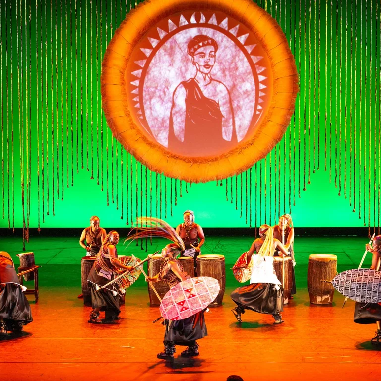 Mehrere Performerinnen tanzen mit Schilden und Speeren vor einer Reihe von Trommeln. Eine große runde Projektion dominiert den Bühnenhintergrund.