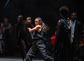 A performer dressed in black moves dynamically across the stage, while other performers stand in the background and the audience surrounds the scene.