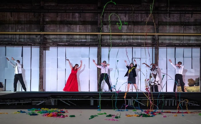Several performers strike expressive poses on a brightly lit, elevated stage in front of a white panel wall inside an old industrial hall. In the foreground, colorful paper streamers float through the air.