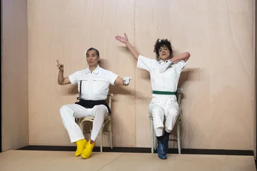 Marlene Monteiro Freitas und Israel Galván sitzen auf Stühlen vor einer hellen Holzwand und gestikulieren mit erhobenen Armen.