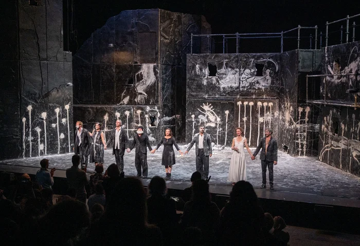 Curtain call from Falaise: The cast stands side by side in front of the dark, white-splattered stage backdrop, holding hands and bowing to the audience.