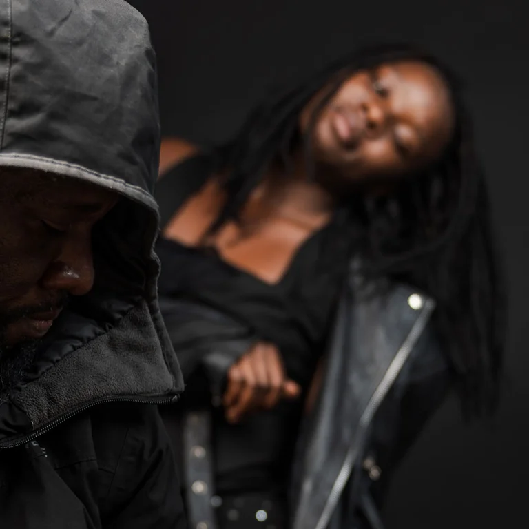 Eine Person sitzt mit gesenktem Blick und hält den Hals einer akustischen Gitarre, während im Hintergrund eine zweite Person in dunkler Kleidung mit geneigtem Kopf eine ruhige, tänzerische Bewegung ausführt.