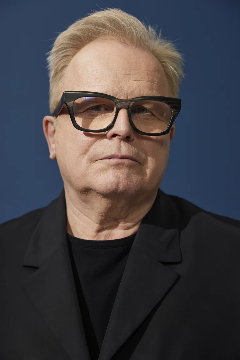 A man with glasses and a black jacket, the artist Herbert Grönemeyer, in front of a blue background