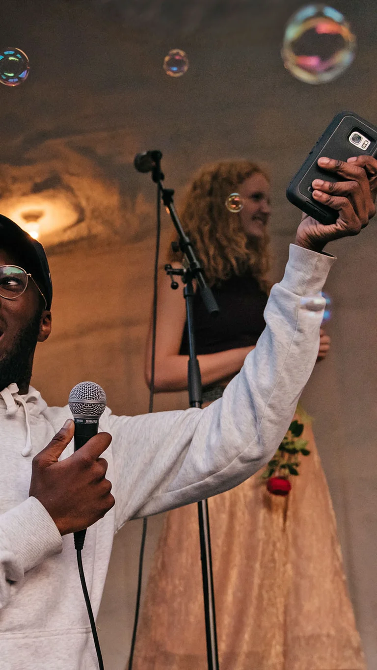 Eine Person hält ein Mikrofon in der einen, sein Smartphone in der anderen Hand, während im Hintergrund weitere junge Menschen auf einer Bühne singen; Seifenblasen schweben durch den Raum.