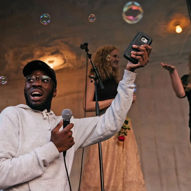 Eine Person hält ein Mikrofon in der einen, sein Smartphone in der anderen Hand, während im Hintergrund weitere junge Menschen auf einer Bühne singen; Seifenblasen schweben durch den Raum.