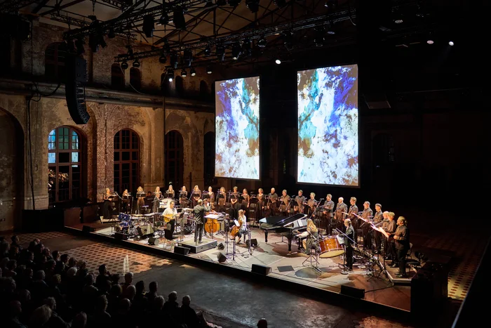 Konzert mit Chorwerk Ruhr und Bang on a Can All-Stars auf einer Bühne in einer Industriehalle, im Hintergrund zwei große Projektionen mit abstrakten Bildern.