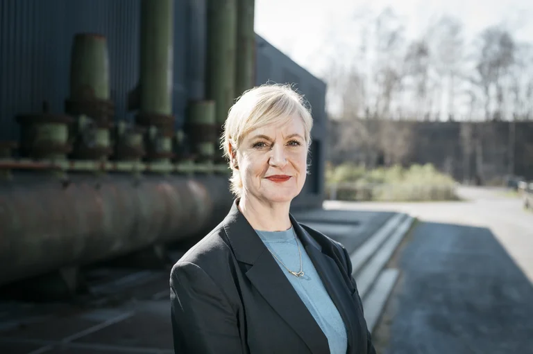Anita van Dolen, curator/consultant for dance, stands in front of an industrial backdrop and looks into the camera.