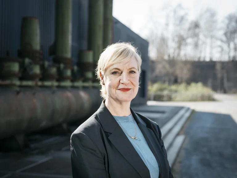 Anita van Dolen, Kuratorin/ Beraterin Bereich Tanz. steht vor Industriekulisse und blickt in die Kamera.