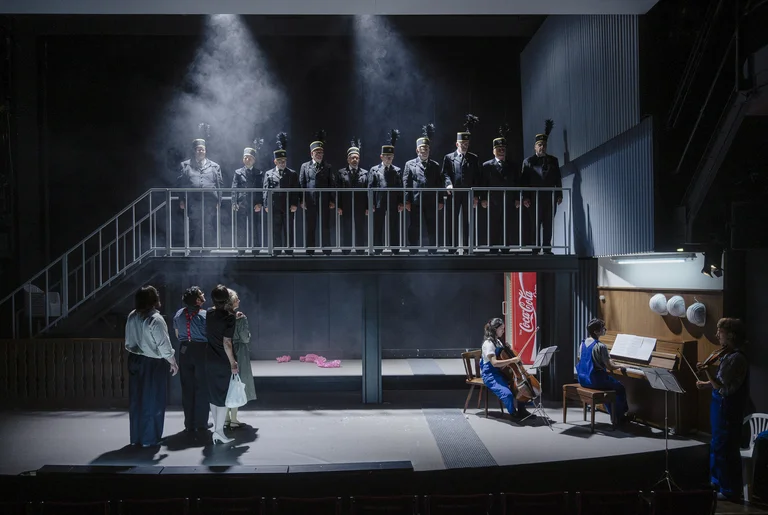 A stage scene with ten people in black miners’ uniforms standing on a raised platform. Four other people stand below, while three musicians on the right play cello, piano and violin.