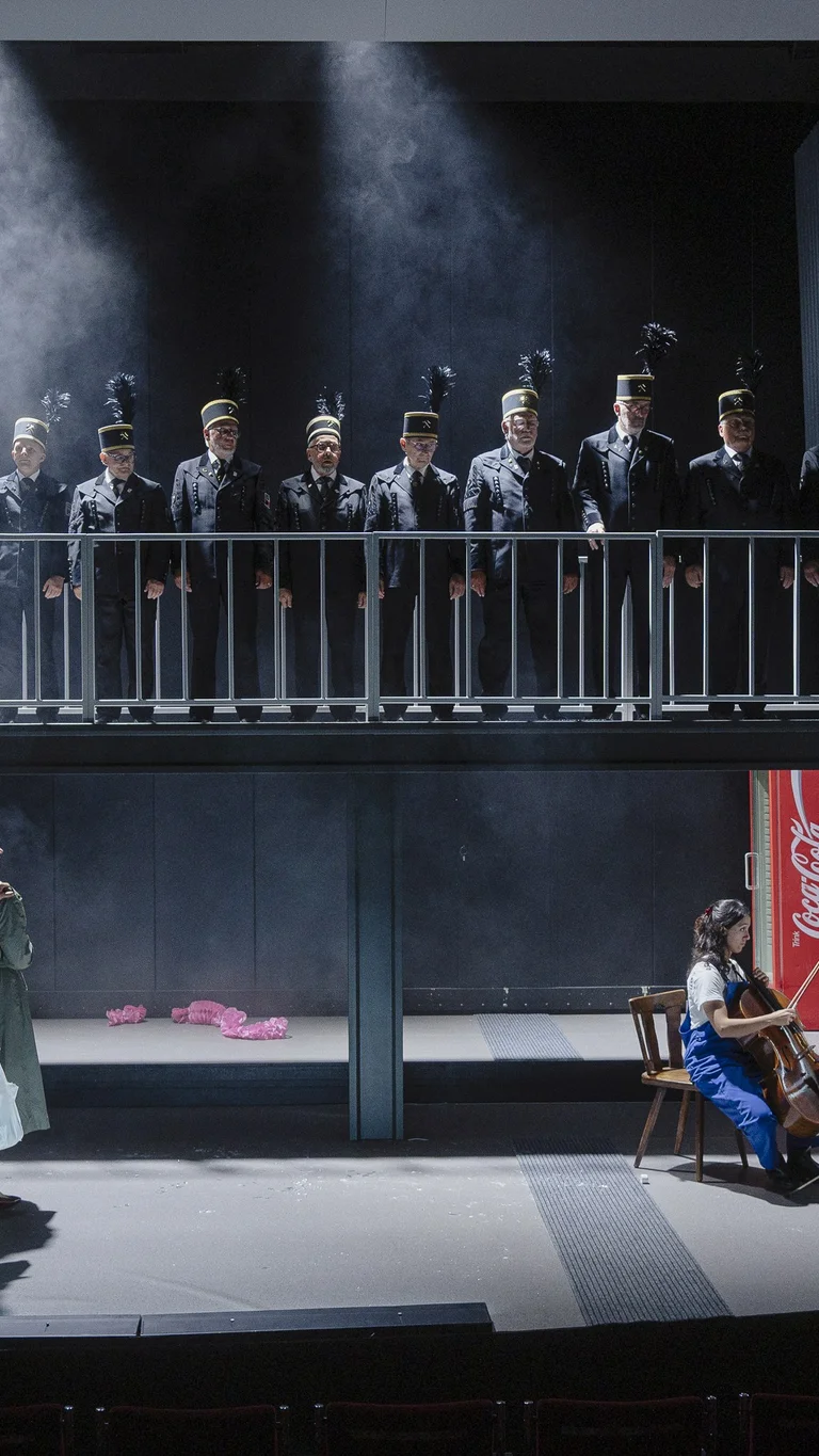 A stage scene with ten people in black miners’ uniforms standing on a raised platform. Four other people stand below, while three musicians on the right play cello, piano and violin.