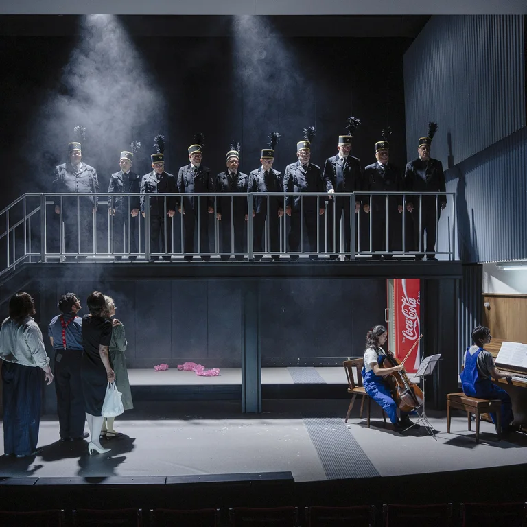 A stage scene with ten people in black miners’ uniforms standing on a raised platform. Four other people stand below, while three musicians on the right play cello, piano and violin.