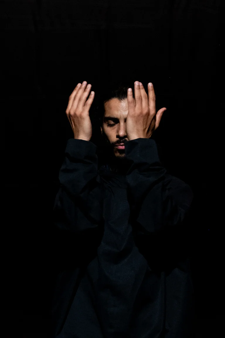 Mohamed Toukabri in dunkler Kleidung steht vor schwarzem Hintergrund mit geschlossenen Augen. Er hebt beide Hände vor das Gesicht, die Handflächen seinem Gesicht zugewandt, in einer konzentrierten, fast meditativen Pose.