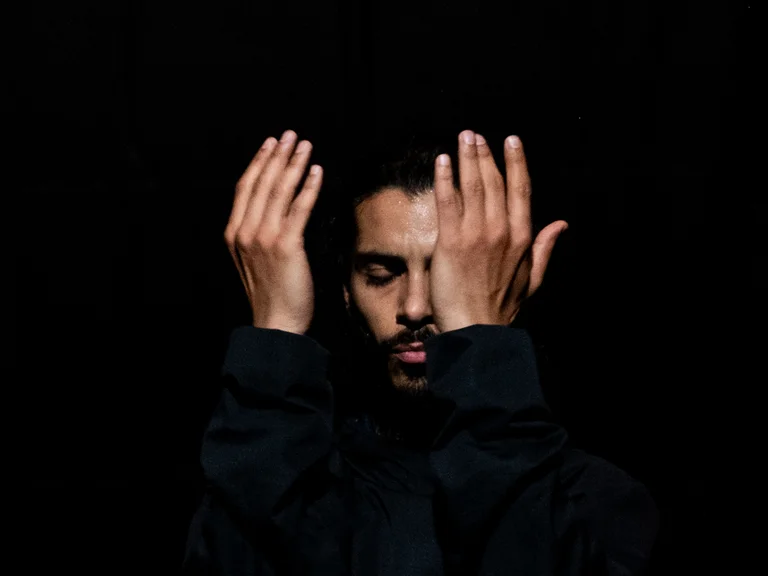 Mohamed Toukabri in dunkler Kleidung steht vor schwarzem Hintergrund mit geschlossenen Augen. Er hebt beide Hände vor das Gesicht, die Handflächen seinem Gesicht zugewandt, in einer konzentrierten, fast meditativen Pose.
