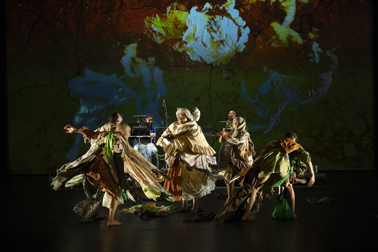 Performers in colorful, flowing costumes dance on stage, with a vibrant projection of abstract floral motifs in the background.